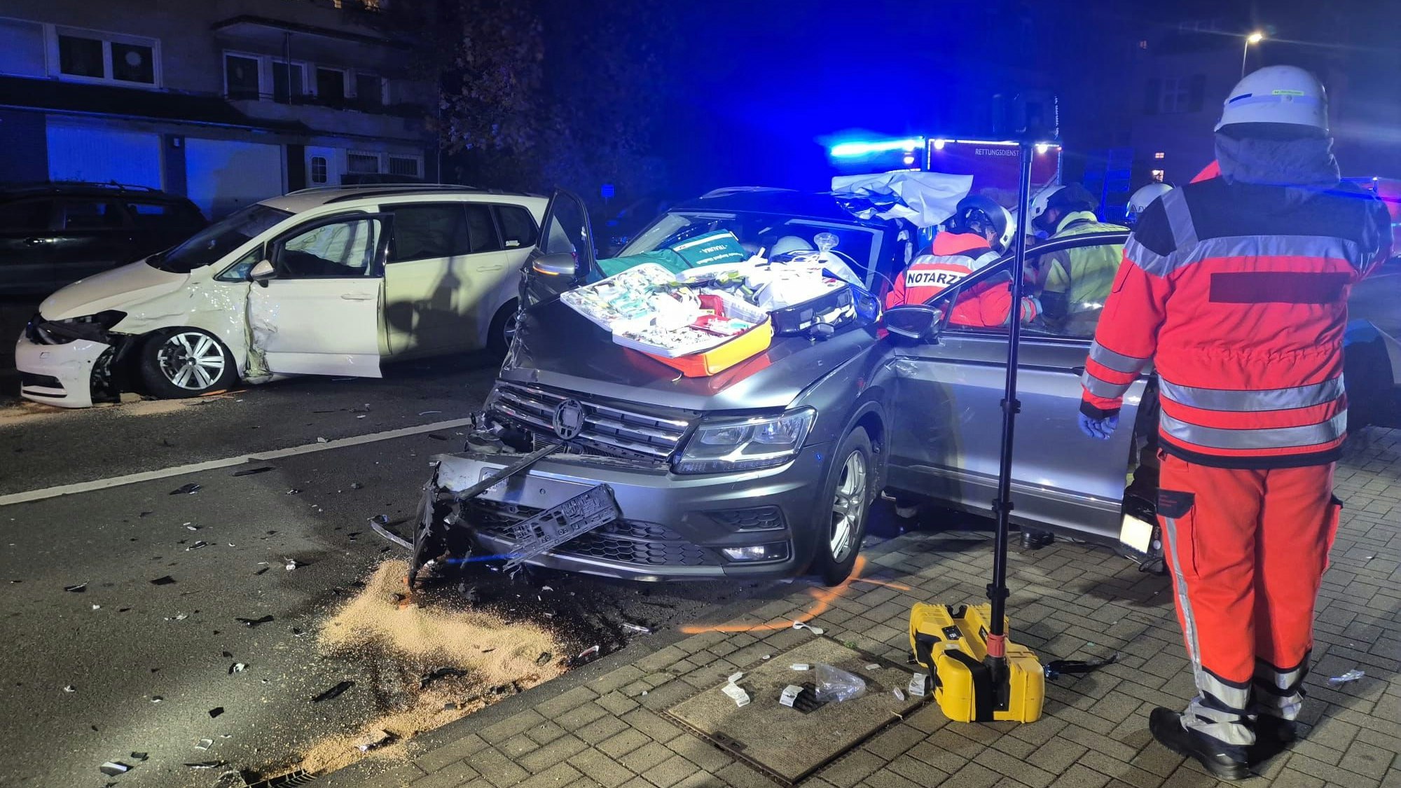 Ein Feuerwehrmann steht vor einem verunfallten Auto, die Fahrertür steht offen, dort kümmert sich unter anderem ein Notarzt um einen Insassen.