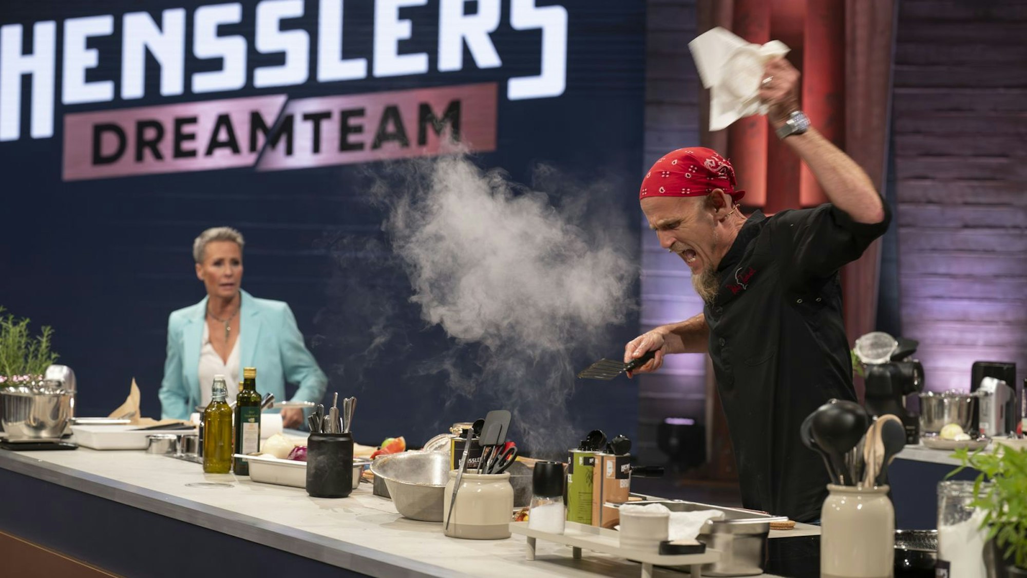 Ein Mann steht am Herd und reißt einen Arm hoch, als Qualm aufsteigt. Ein paar Meter neben ihm steht eine Frau.