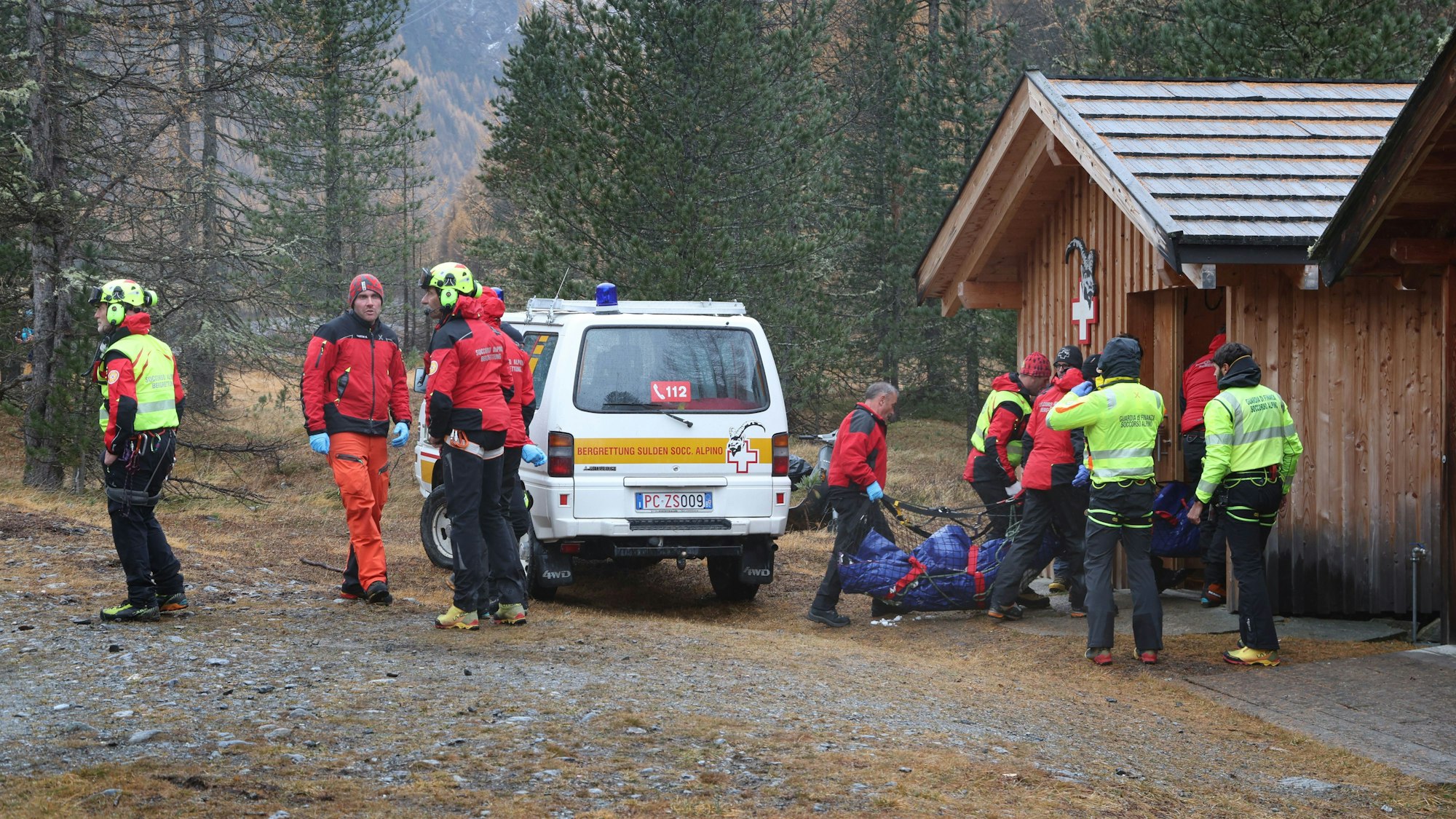 Mehrere Einsatzkräfte stehen vor einer Holzhütte, zwei von ihnen tragen einen Leichensack zur Hütte.