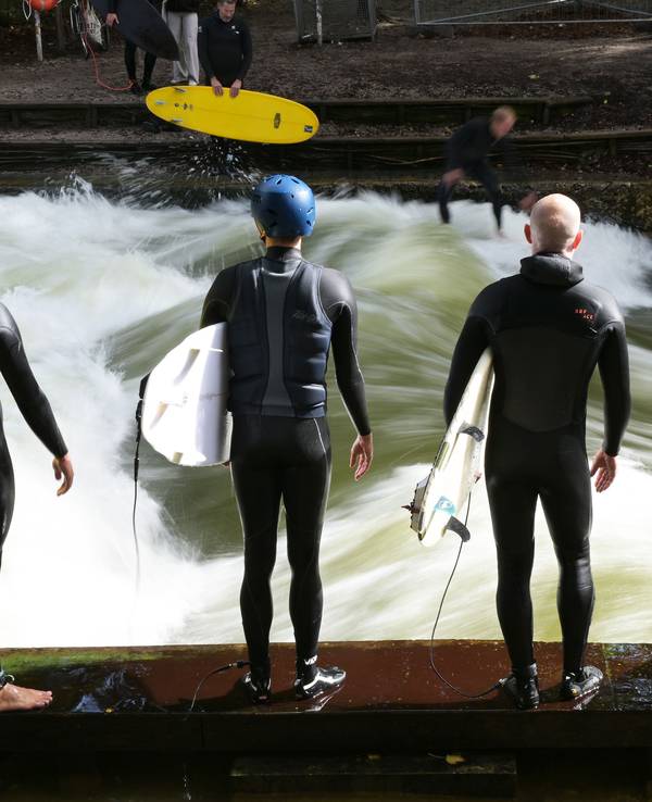 Vier Männer warten mit Surfbrettern an der Eisbachwelle im Englischen Garten.