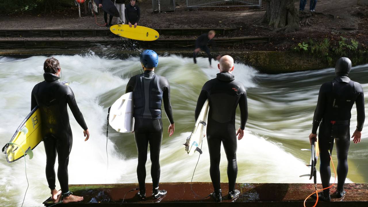 Vier Männer warten mit Surfbrettern an der Eisbachwelle im Englischen Garten.