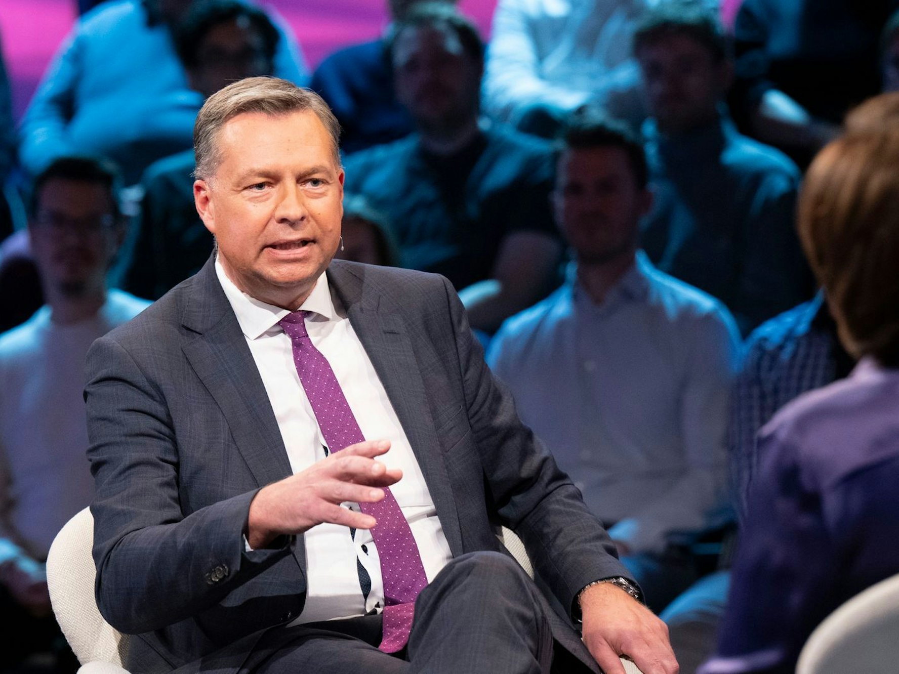 Ein Mann gestikuliert auf einem Stuhl sitzend, hinter ihm sitzt das Publikum.