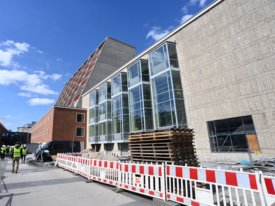 Seitlich an einem Gebäude steht ein Bauzaun, dahinter sind Baumaterialien gelagert.