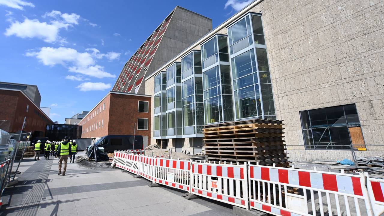Seitlich an einem Gebäude steht ein Bauzaun, dahinter sind Baumaterialien gelagert.