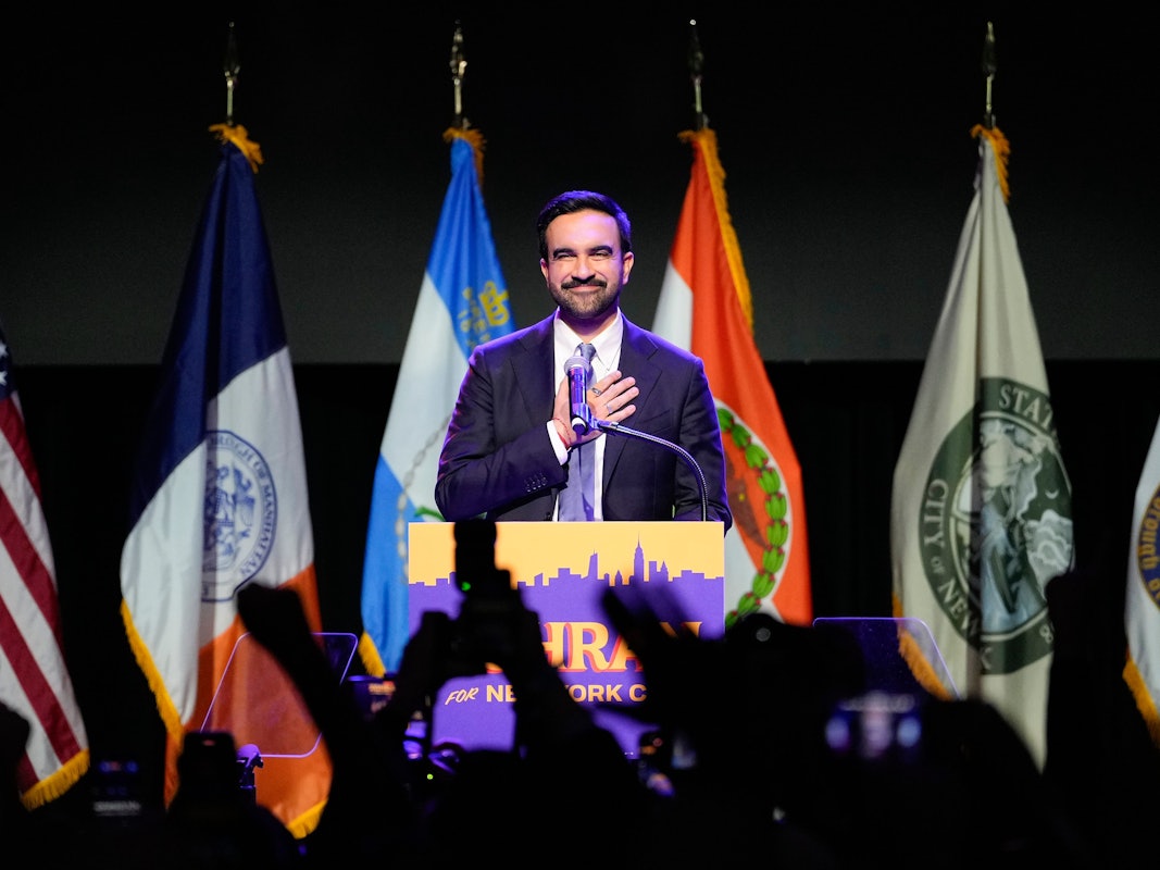 Ein Mann steht an einem Rednerpult und hält seine rechte Hand auf seinen Oberkörper, im Hintergrund stehen Fahnen, im Vordergrund sind in die Höhe gestreckte Hände zu sehen.