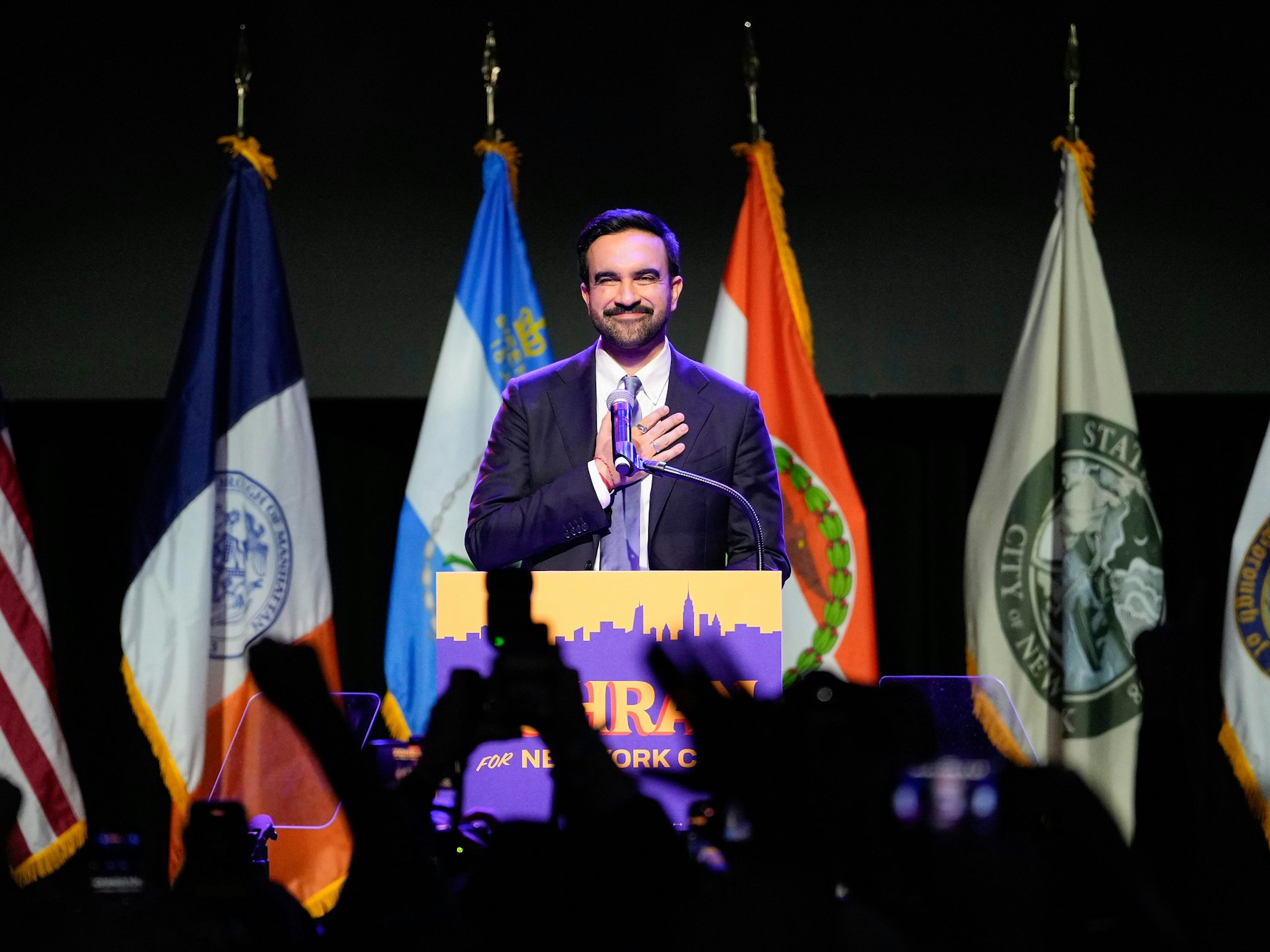 Ein Mann steht an einem Rednerpult und hält seine rechte Hand auf seinen Oberkörper, im Hintergrund stehen Fahnen, im Vordergrund sind in die Höhe gestreckte Hände zu sehen.