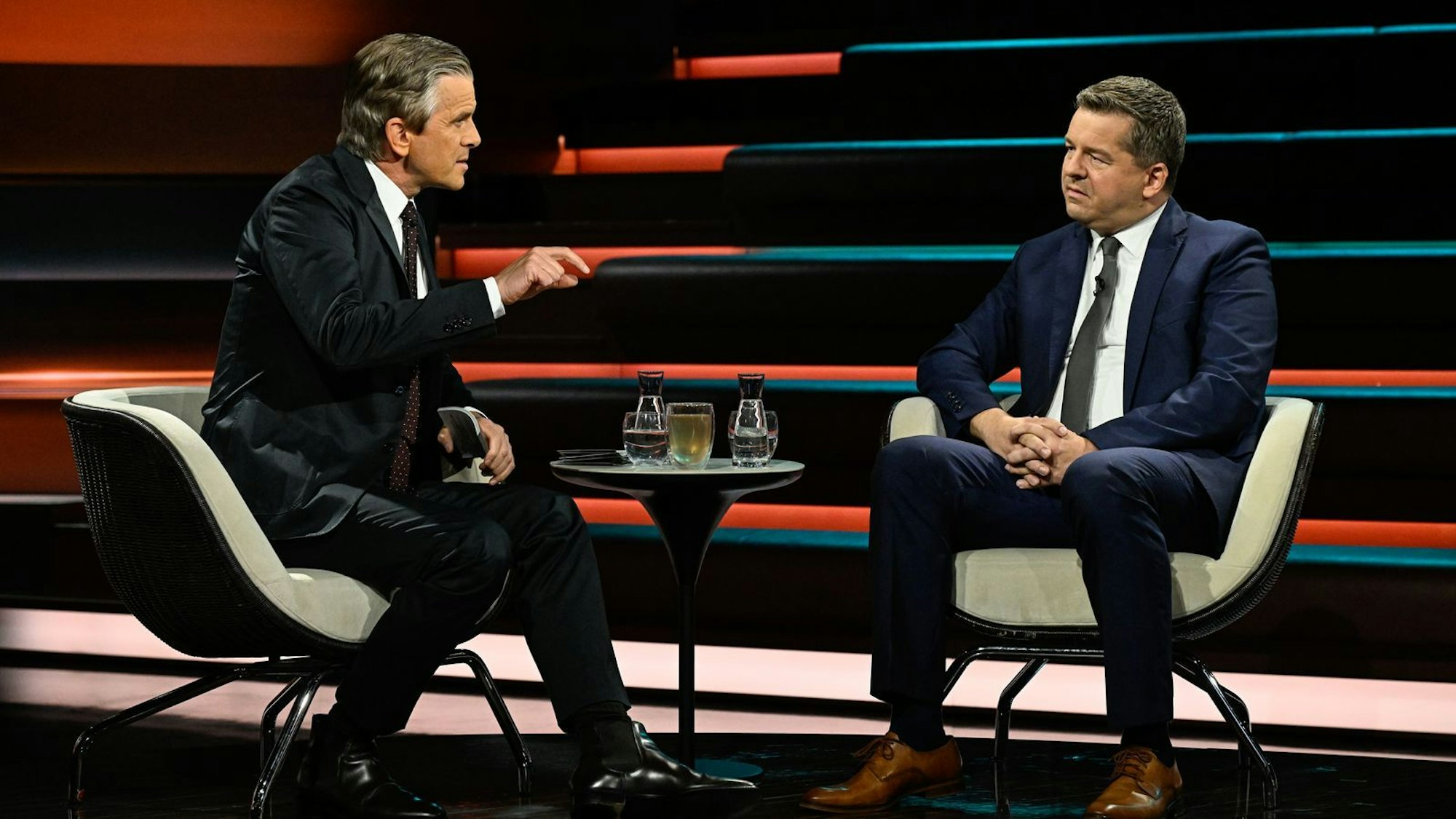 Zwei Männer sitzen in Sesseln in einem TV-Studio und gucken sich an.