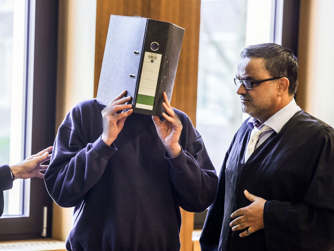 Der Angeklagte Krankenpfleger (M) steht im November 2025 mit einem Aktenordner vor dem Gesicht neben seinem Anwalt Volker Breyer (r) im Gerichtssaal des Aachener Landgerichts.