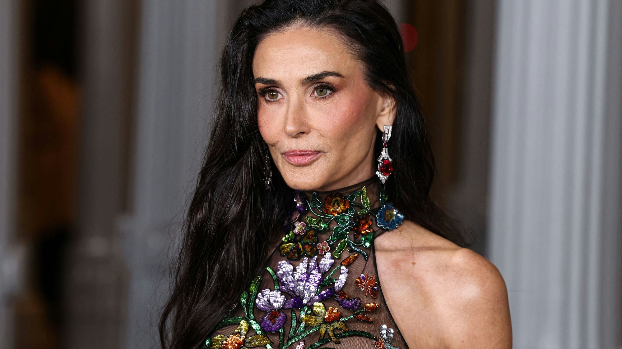 Demi Moore mit langen, dunklen Haaren, besticktem Kleid und roten Juwelenohrringen.