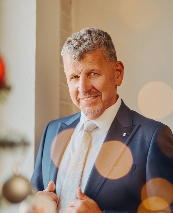 Ein Mann mit graumelierten Locken, blauem Anzug und weißem Hemd lächelt milde in die Kamera.