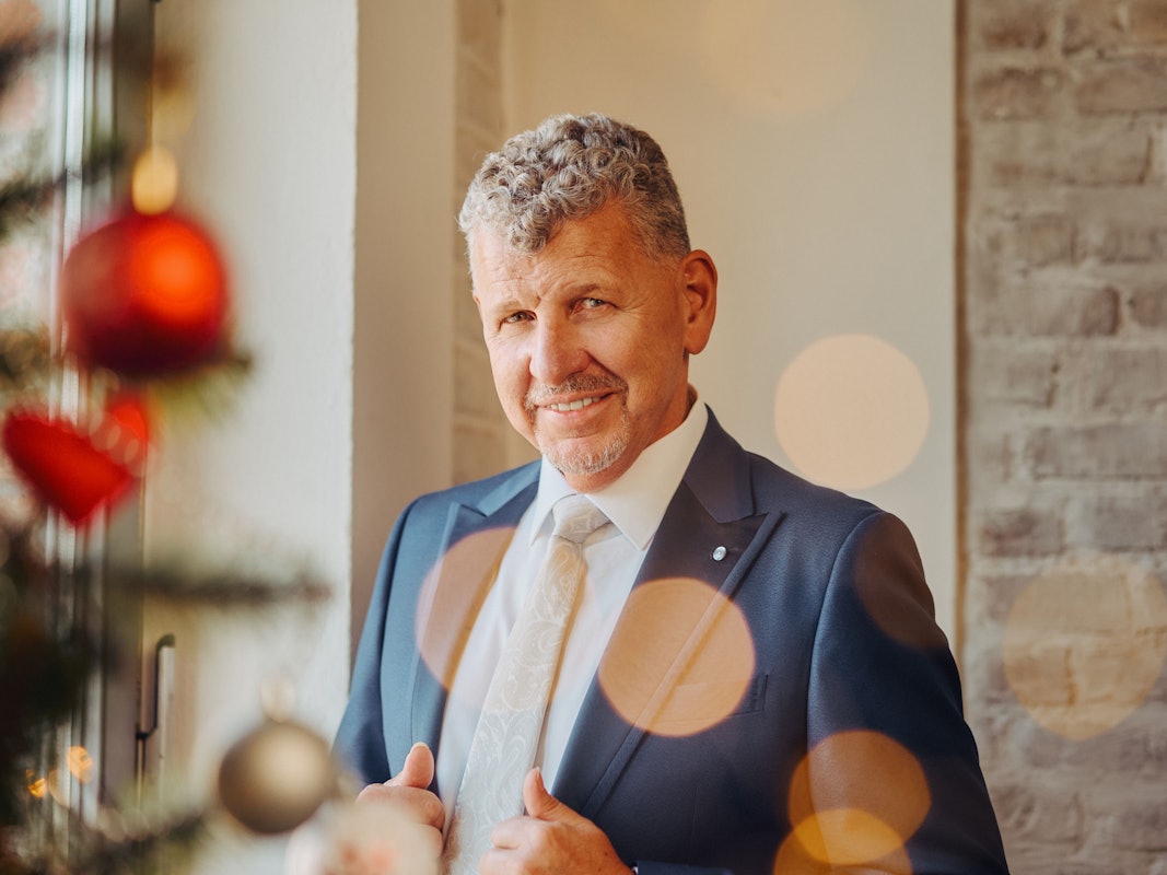 Ein Mann mit graumelierten Locken, blauem Anzug und weißem Hemd lächelt milde in die Kamera.