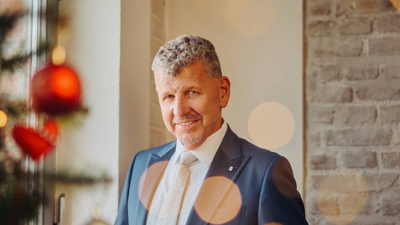 Ein Mann mit graumelierten Locken, blauem Anzug und weißem Hemd lächelt milde in die Kamera.