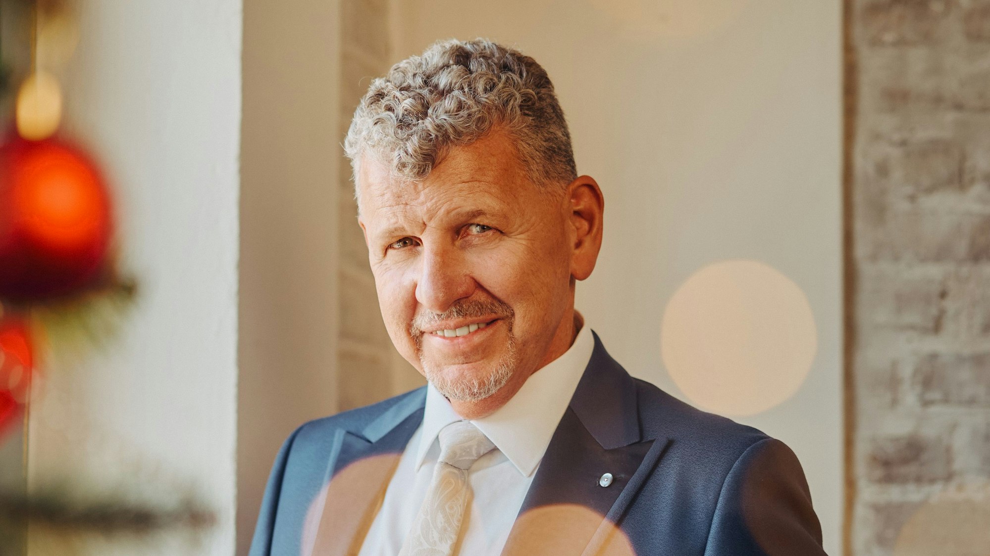 Ein Mann mit graumelierten Locken, blauem Anzug und weißem Hemd lächelt milde in die Kamera.