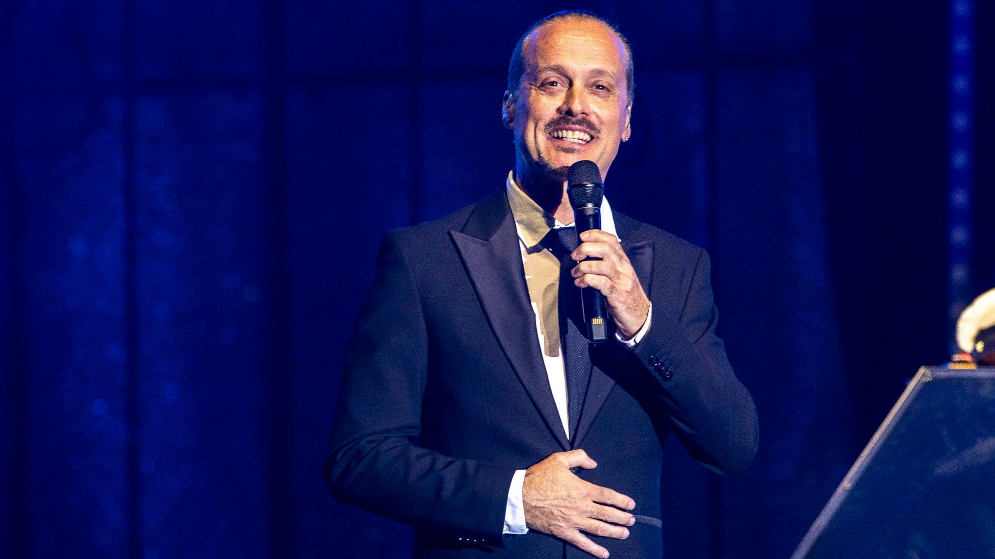 Ein Mann im blauen Anzug steht vor einer blauen Wand und spricht etwas in ein Mikrofon.