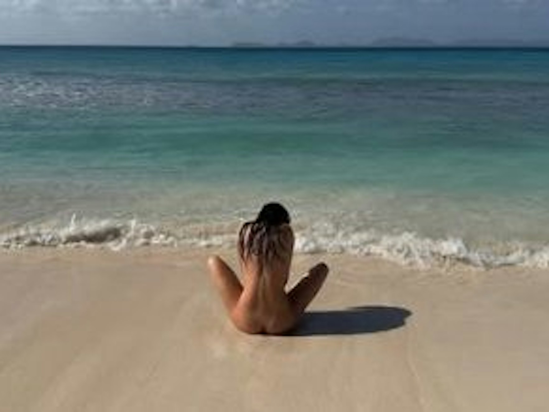 Eine Frau sitzt nackt an einem Strand, man sieht sie von hinten.