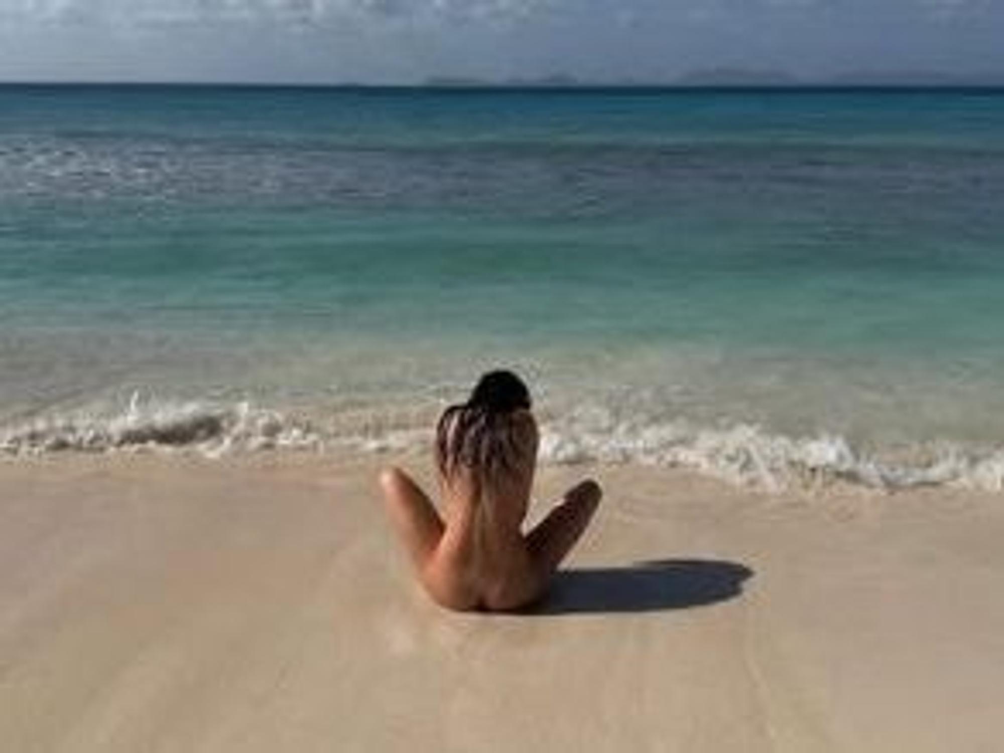 Eine Frau sitzt nackt an einem Strand, man sieht sie von hinten.