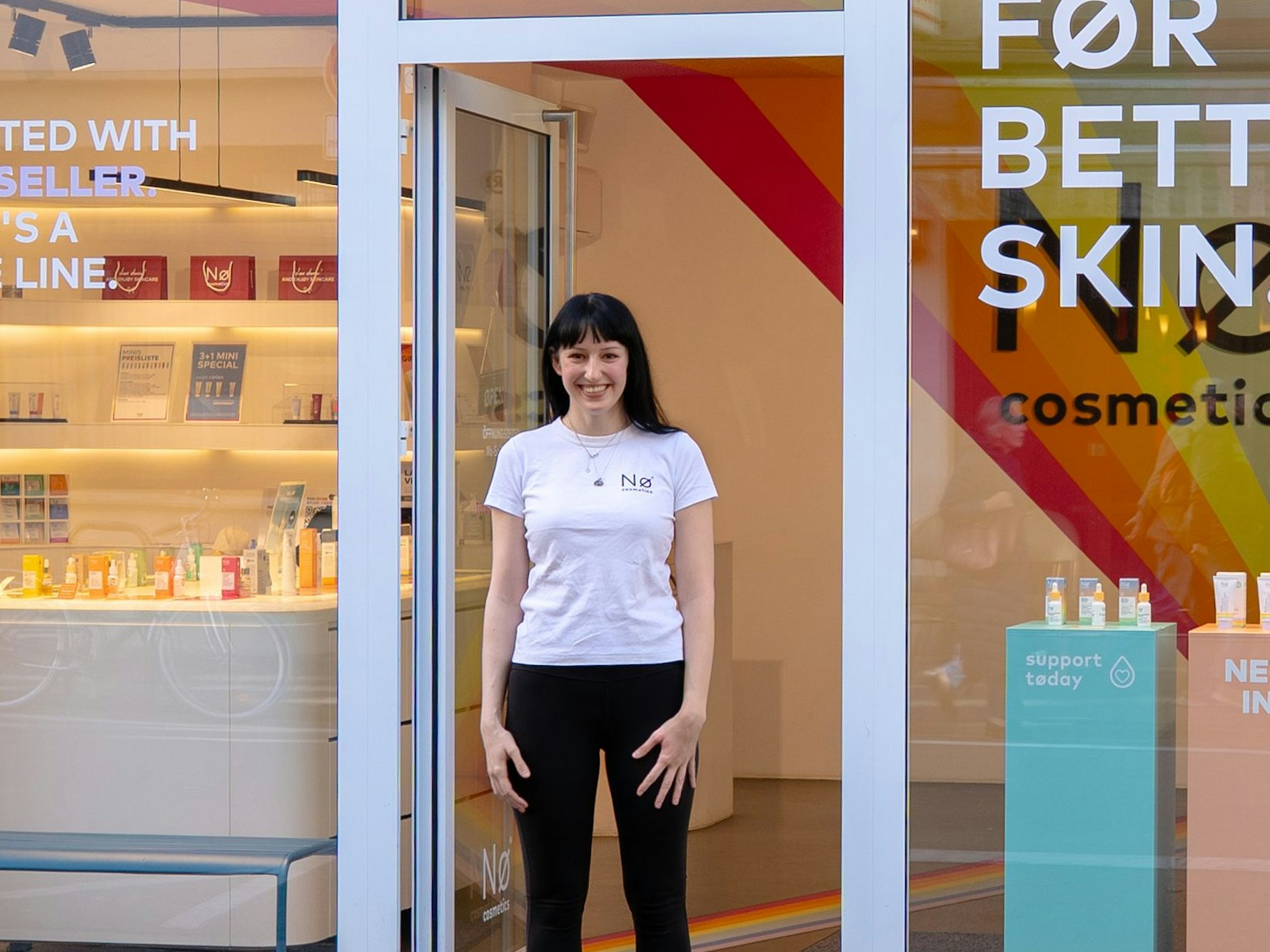 Eine junge Frau in weißem T-Shirt steht in der Tür eines Kosmetikgeschäftes und lächelt.