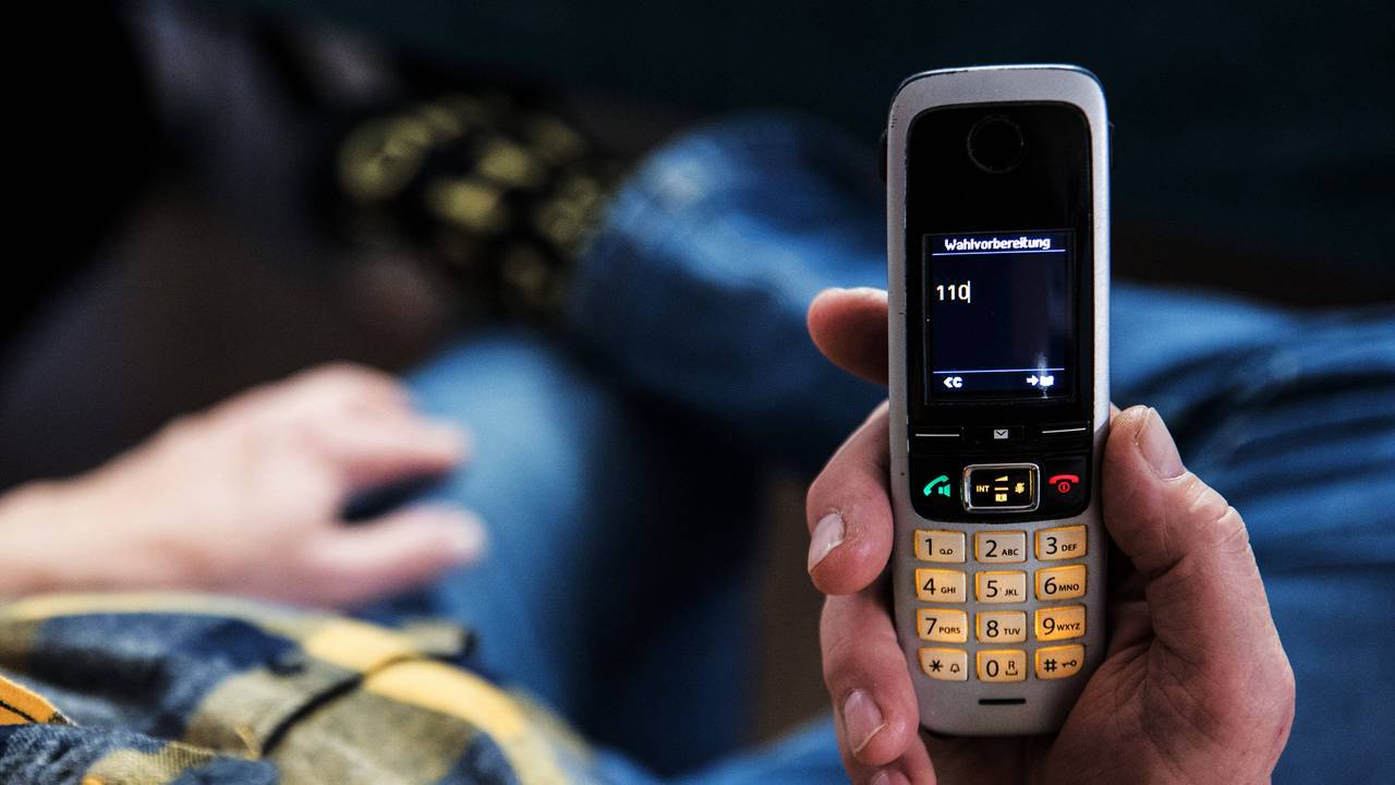 Ein älterer Menschen sitzt in seiner Wohnung und hält ein Mobiltelefon in der Hand auf dem Display des Telefons ist die 110 zu lesen.