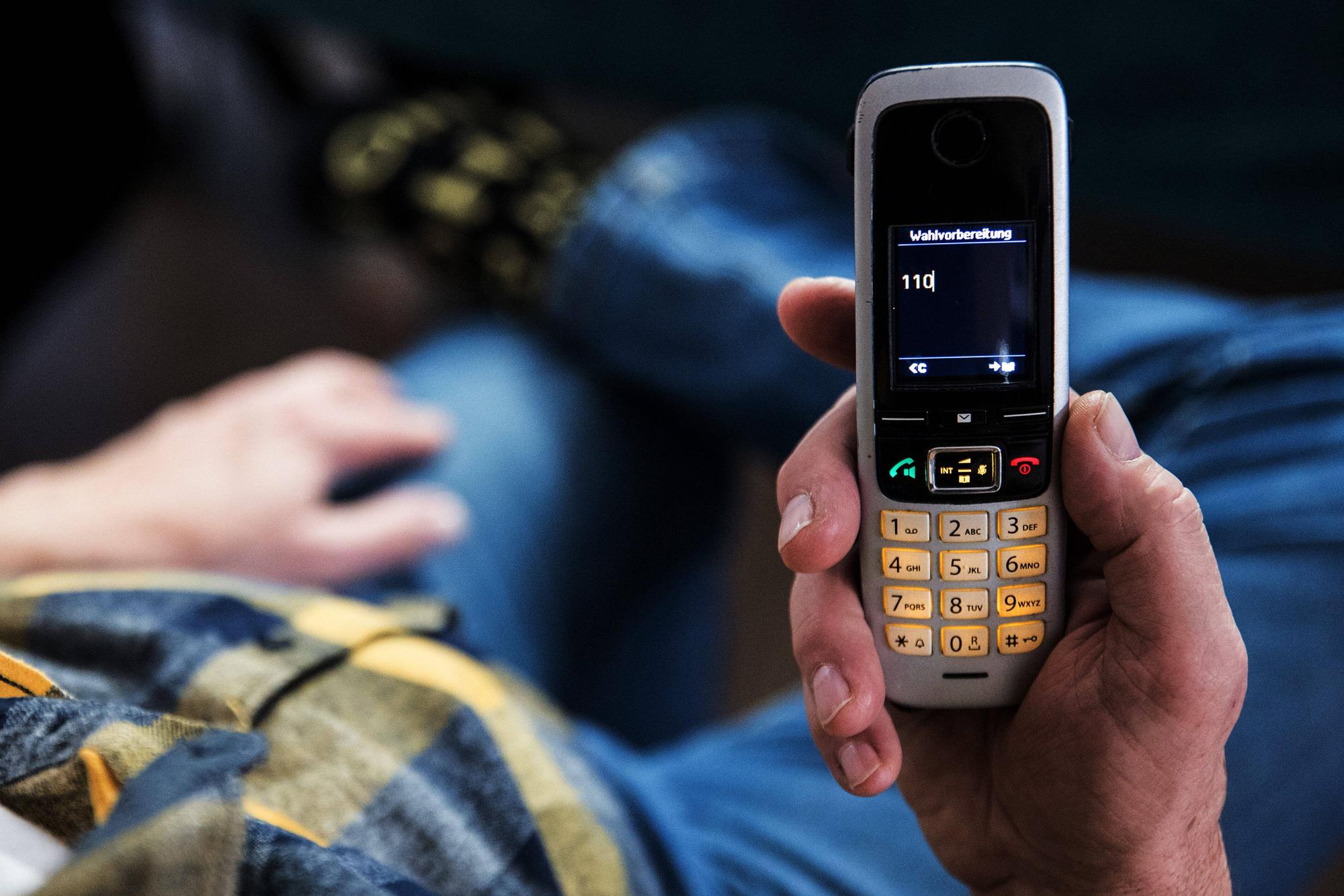 Ein älterer Menschen sitzt in seiner Wohnung und hält ein Mobiltelefon in der Hand auf dem Display des Telefons ist die 110 zu lesen.