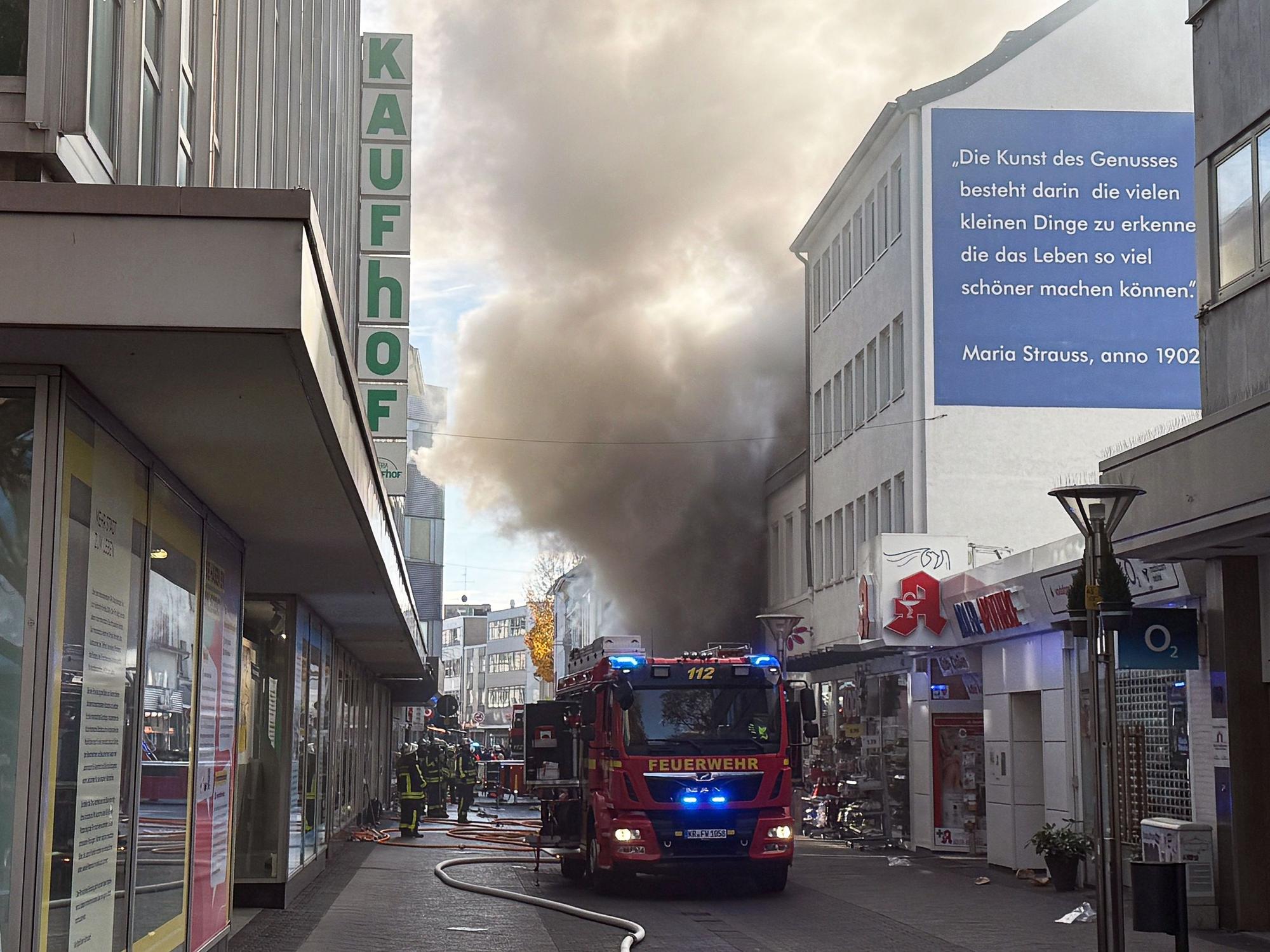 Einsatzkräfte stehen in einer Einkaufstraße vor einem Geschäft, aus dem dichter Rauch quillt. Ein Feuer in einem Laden der Einkaufsmeile Hochstraße hat einen Großeinsatz der Feuerwehr in der Innenstadt ausgelöst.