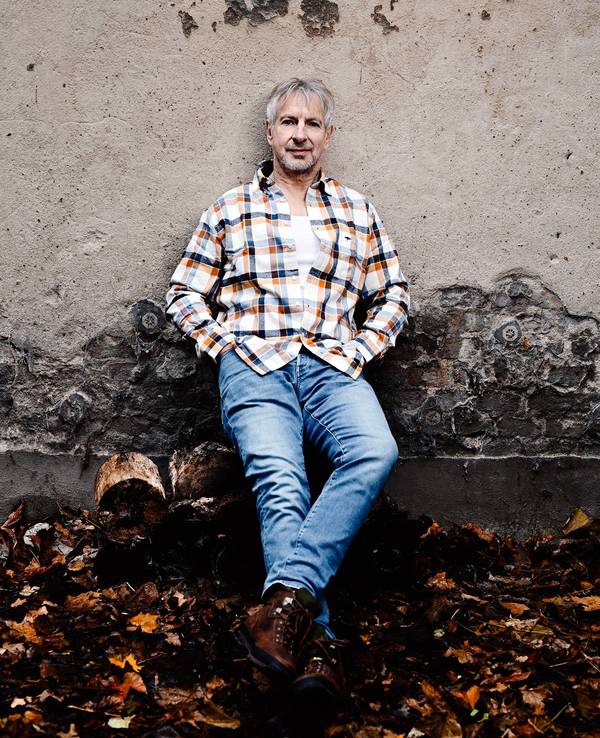 Ein Mann mit grauen Haaren, in Jeans und Karohemd, lehnt lässig an einer Mauer, vor der Herbstlaub liegt.