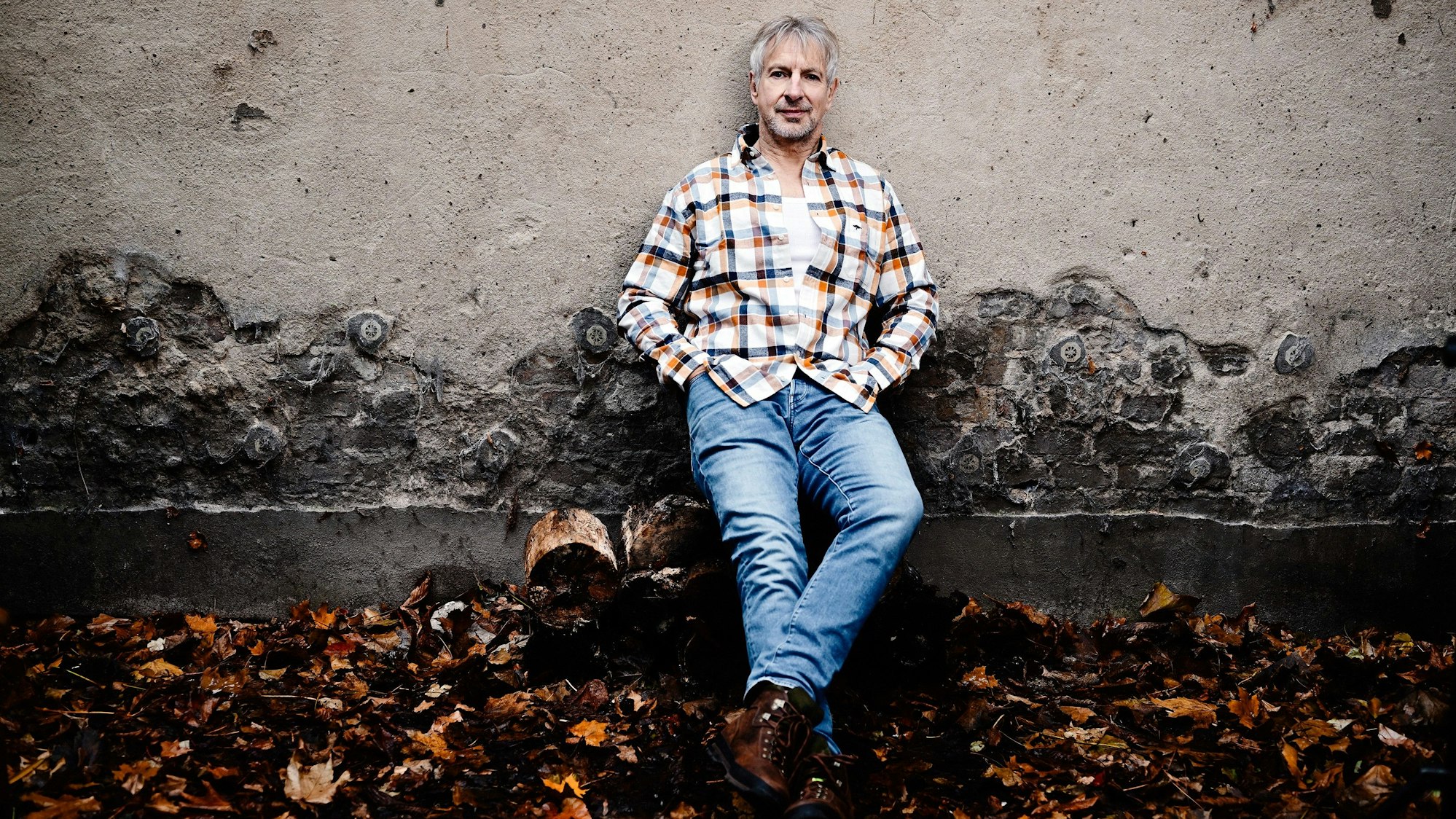 Ein Mann mit grauen Haaren, in Jeans und Karohemd, lehnt lässig an einer Mauer, vor der Herbstlaub liegt.