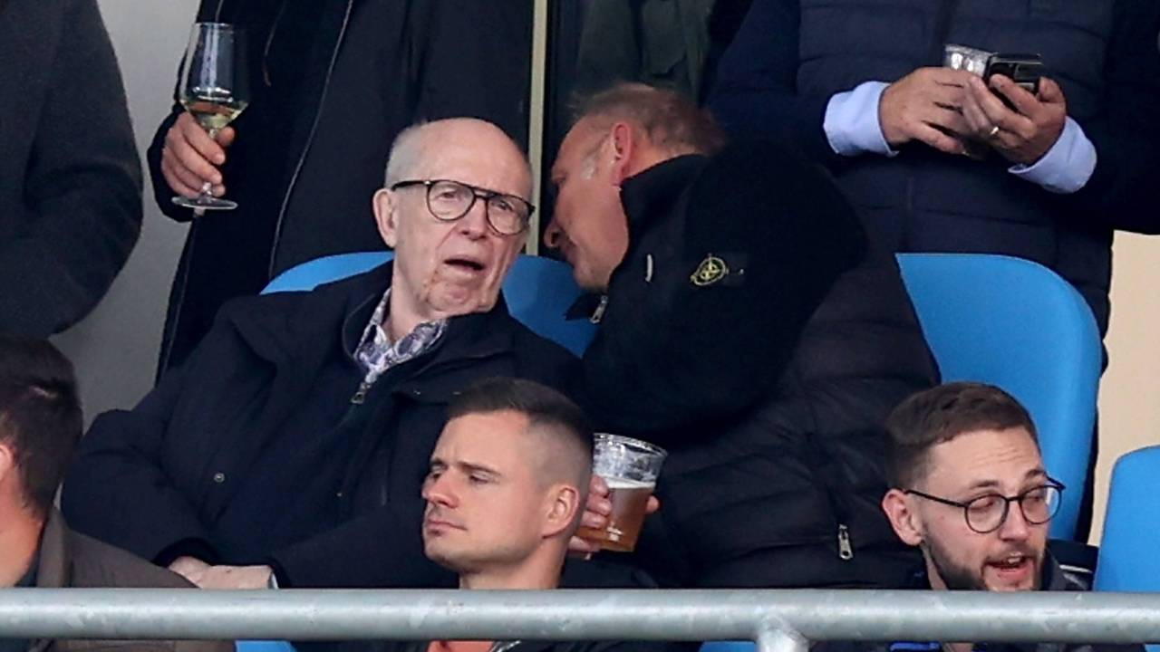 Reiner Calmund sitzt auf der Tribüne und bekommt von seinem Sitznachbarn etwas ins Ohr gesagt.