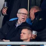 Reiner Calmund sitzt auf der Tribüne und bekommt von seinem Sitznachbarn etwas ins Ohr gesagt.