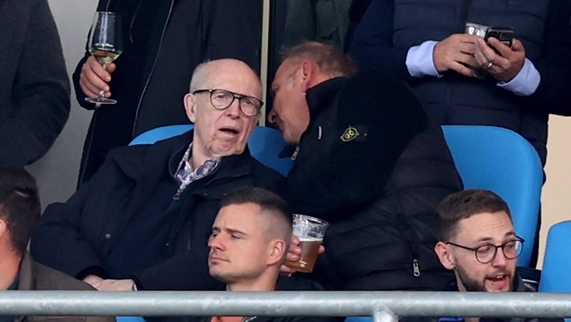 Reiner Calmund sitzt auf der Tribüne und bekommt von seinem Sitznachbarn etwas ins Ohr gesagt.