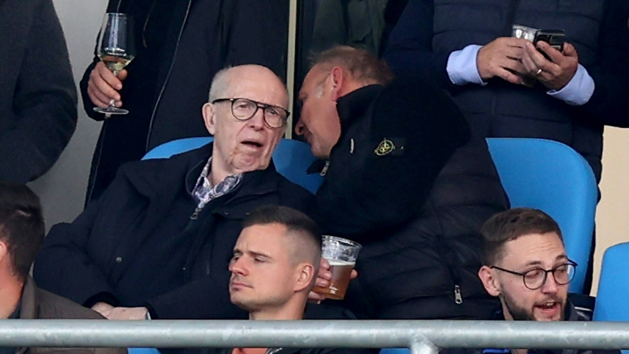 Reiner Calmund sitzt auf der Tribüne und bekommt von seinem Sitznachbarn etwas ins Ohr gesagt.