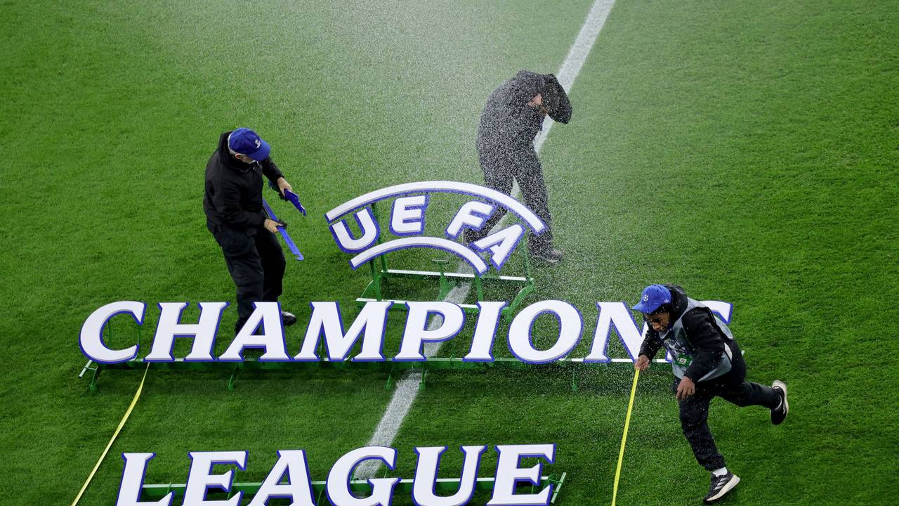 Der Champions League Schriftzug liegt auf dem Rasen, drei Mitarbeiter flüchten vor einem Wasserstrahl.