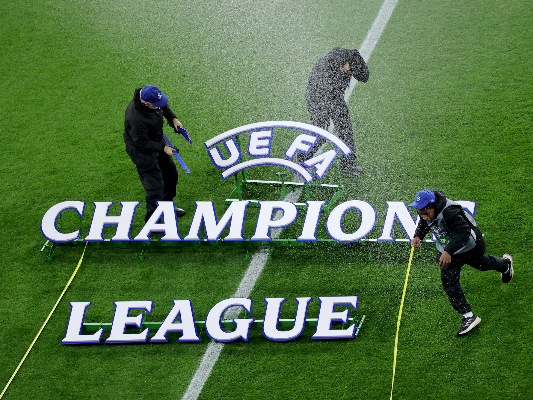 Der Champions League Schriftzug liegt auf dem Rasen, drei Mitarbeiter flüchten vor einem Wasserstrahl.