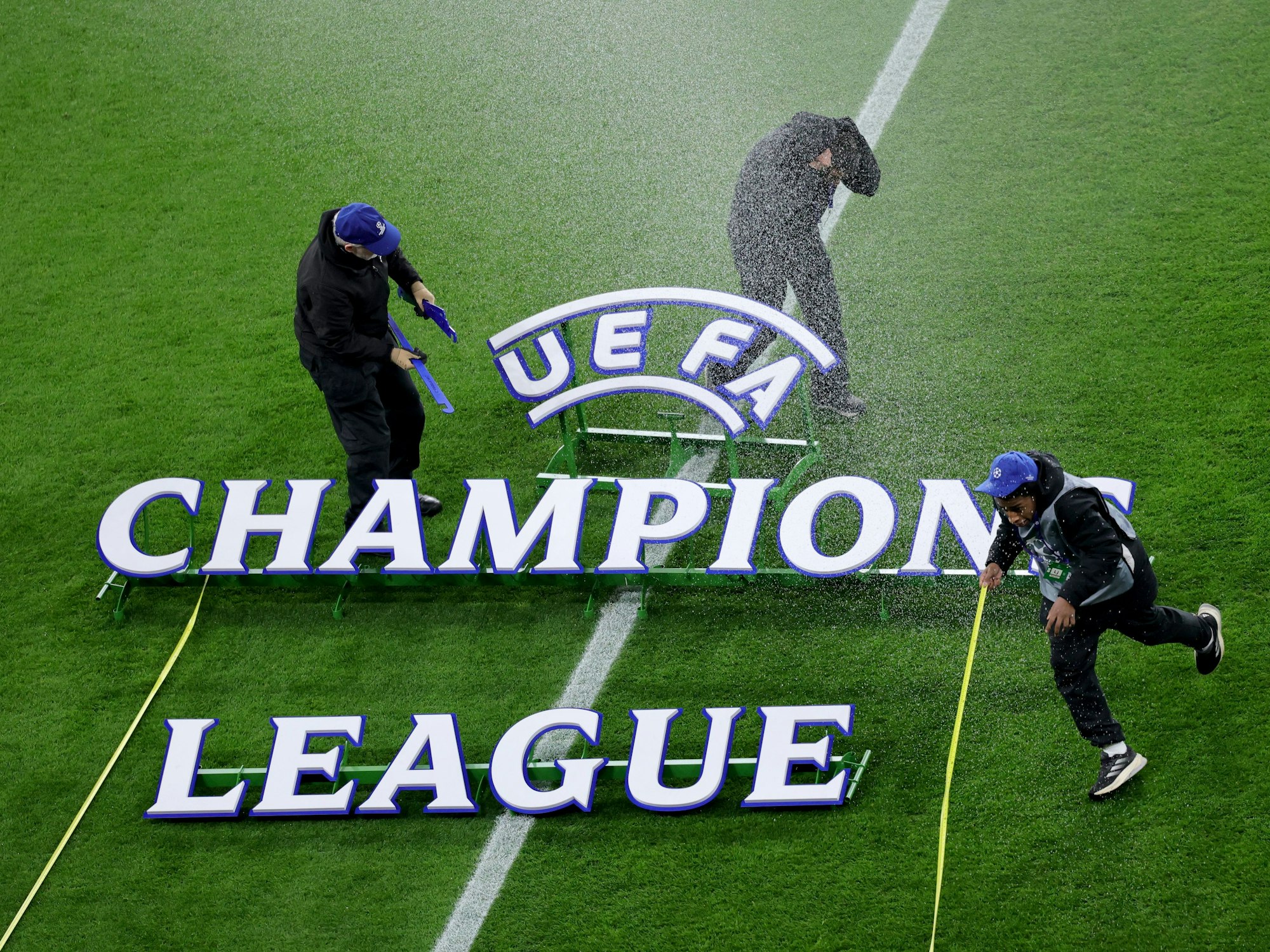 Der Champions League Schriftzug liegt auf dem Rasen, drei Mitarbeiter flüchten vor einem Wasserstrahl.
