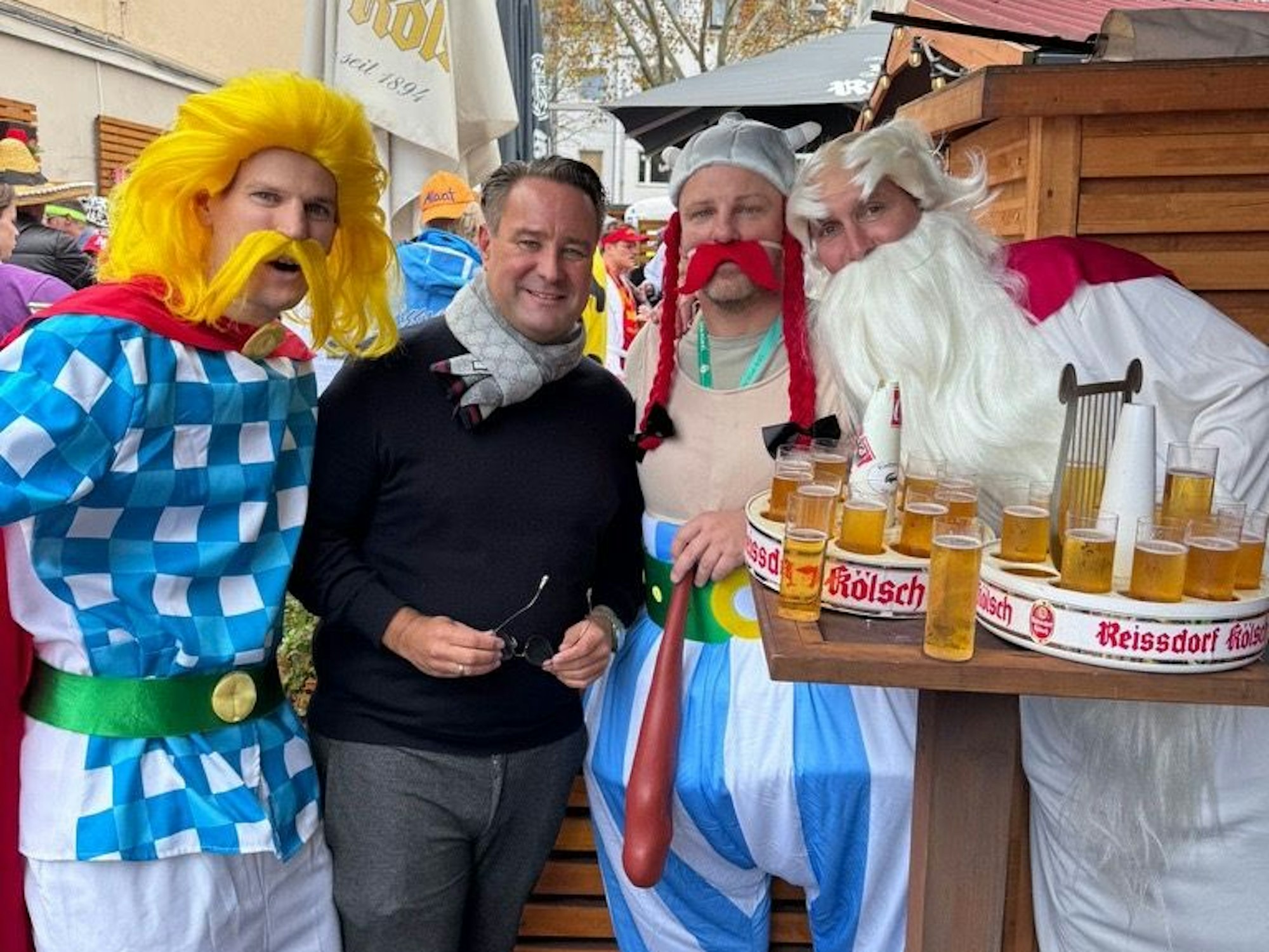 Drei verkleidete Männer, einer mit einem Tablett voller Biergläser, stehen mit einem unverkleideten Mann zusammen.