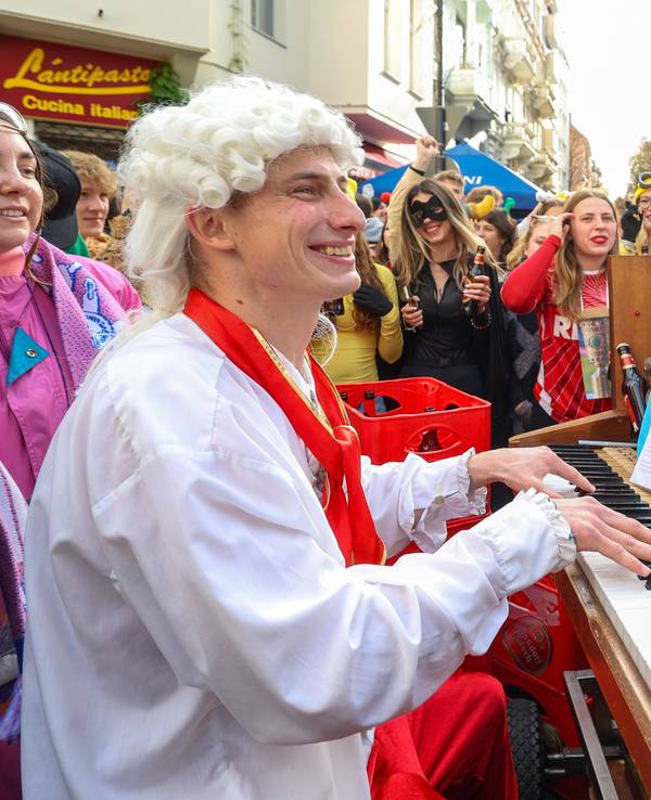 Ein Mann mit Lockenperücke spielt auf einer Straße Klavier und wird von vielen verkleideten Menschen umringt.