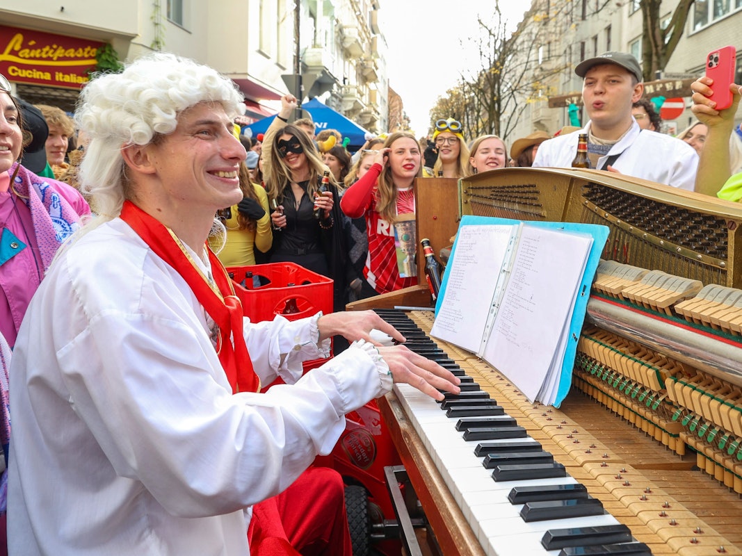 Ein Mann mit Lockenperücke spielt auf einer Straße Klavier und wird von vielen verkleideten Menschen umringt.