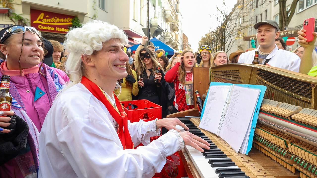 Ein Mann mit Lockenperücke spielt auf einer Straße Klavier und wird von vielen verkleideten Menschen umringt.