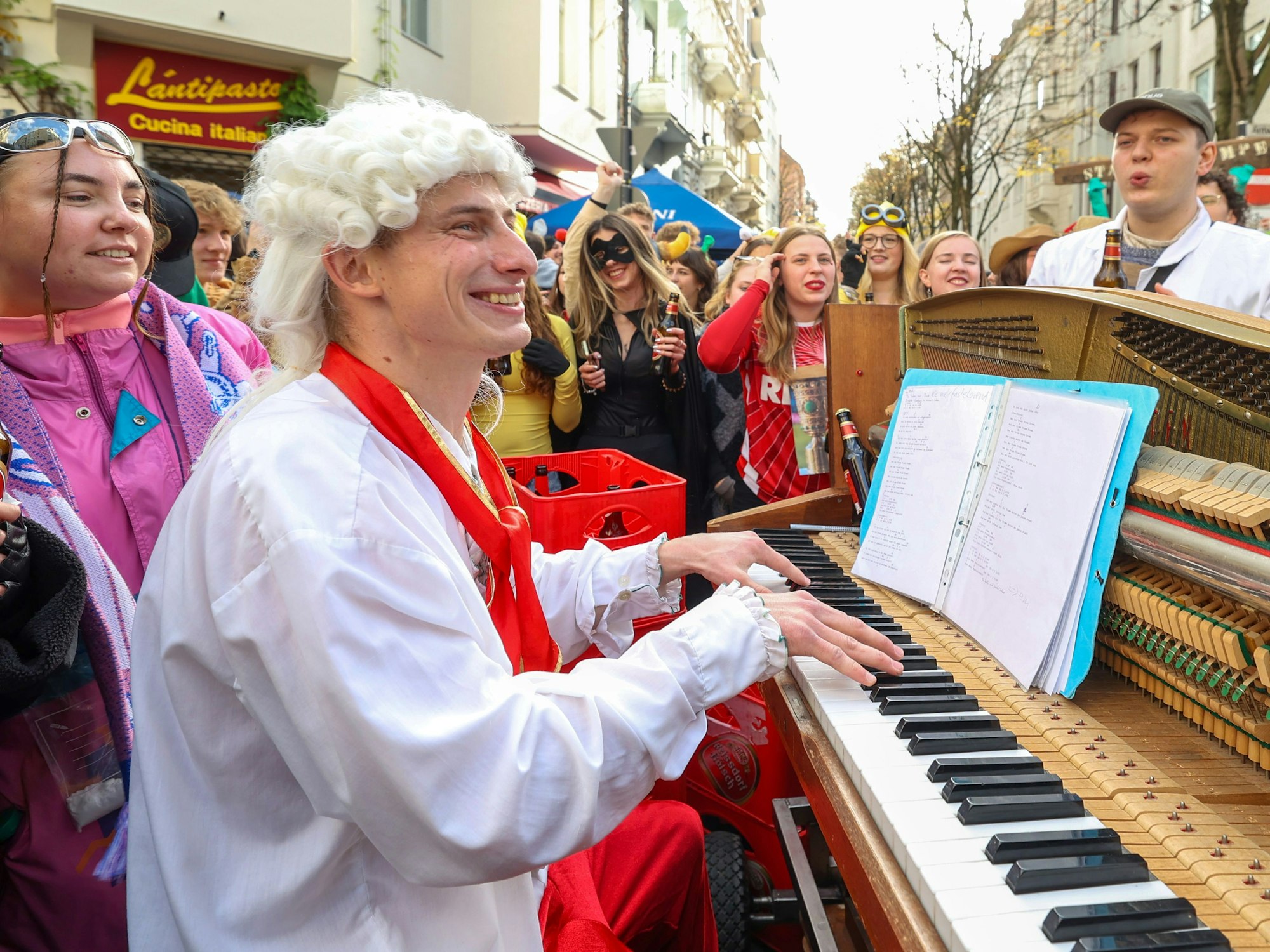 Ein Mann mit Lockenperücke spielt auf einer Straße Klavier und wird von vielen verkleideten Menschen umringt.