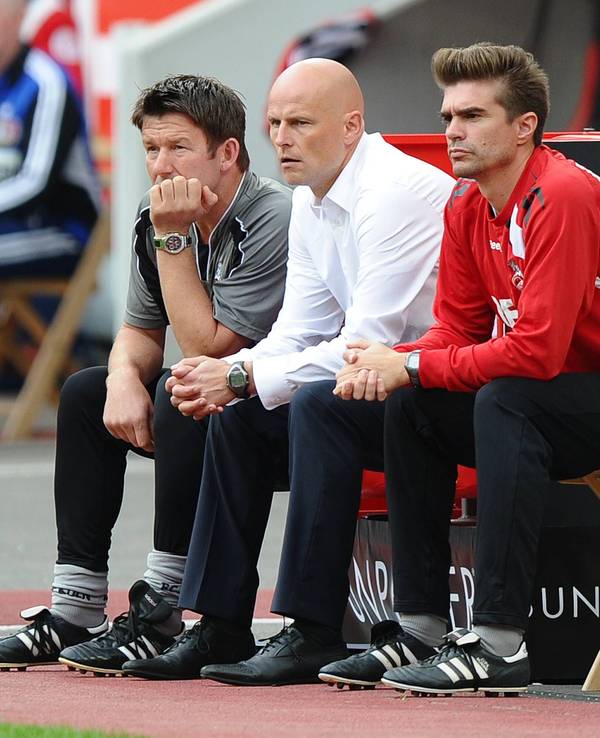 Die Trainer des 1. FC Köln sitzen an der Seitenlinie und blicken aufs Spielfeld.