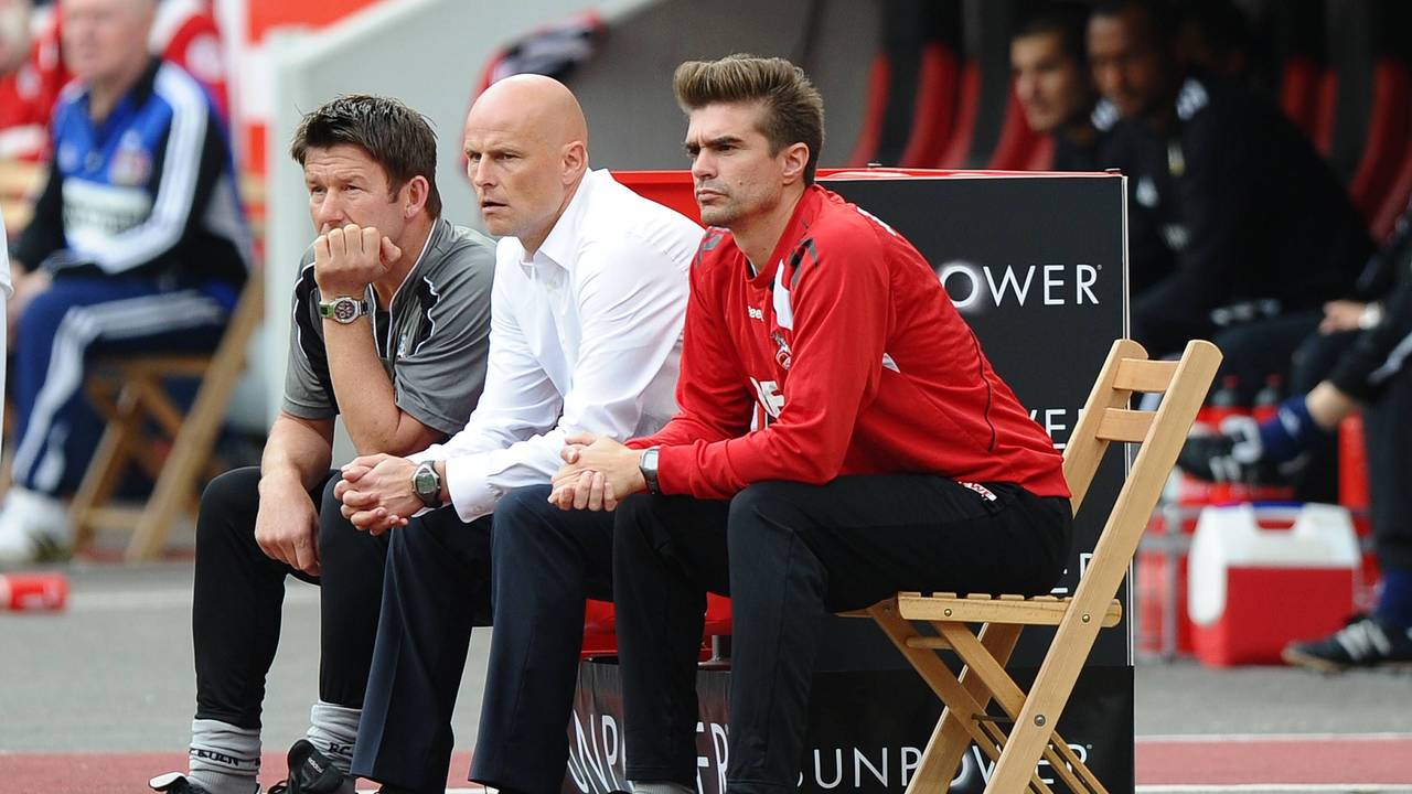Die Trainer des 1. FC Köln sitzen an der Seitenlinie und blicken aufs Spielfeld.