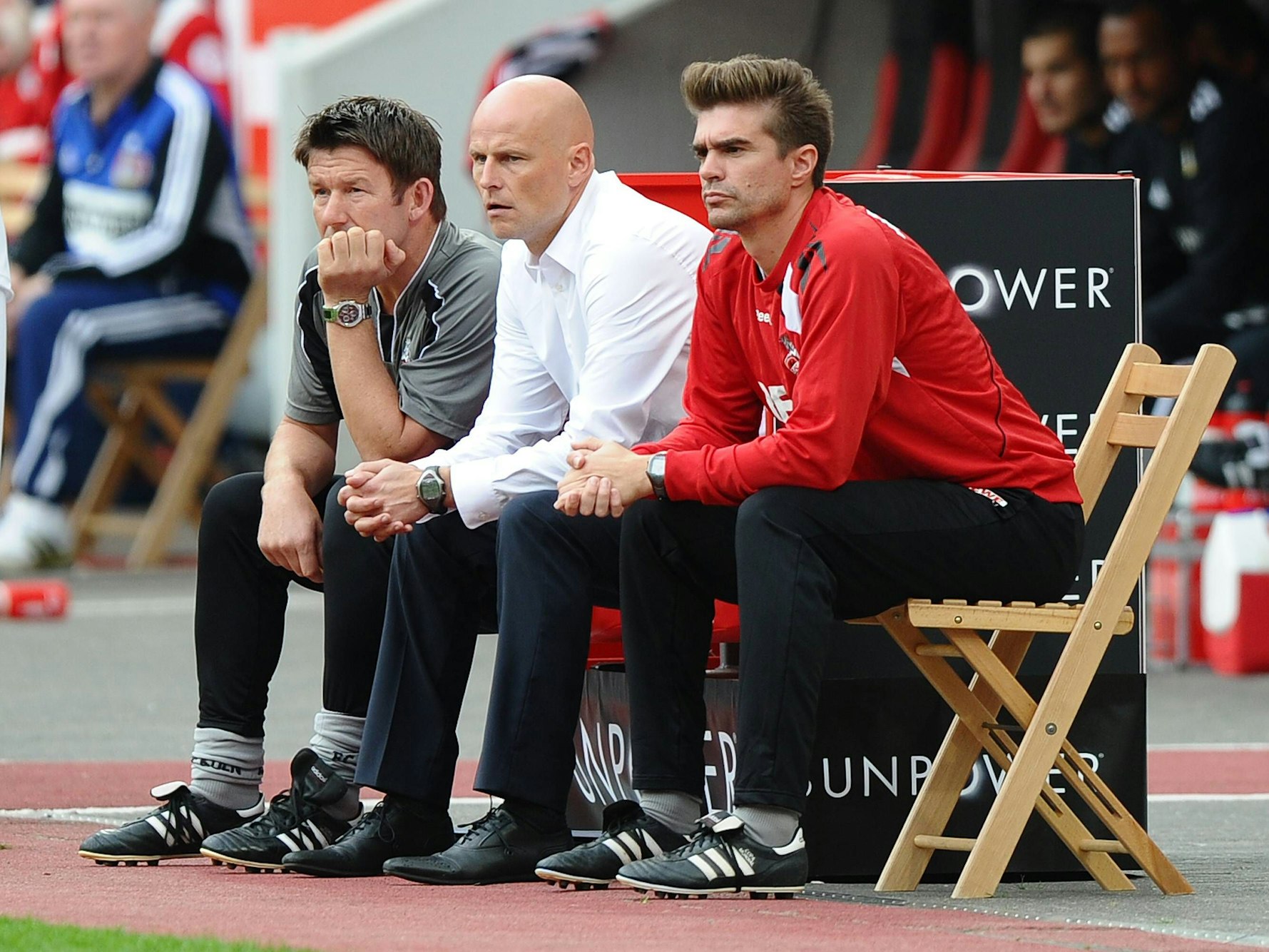 Die Trainer des 1. FC Köln sitzen an der Seitenlinie und blicken aufs Spielfeld.