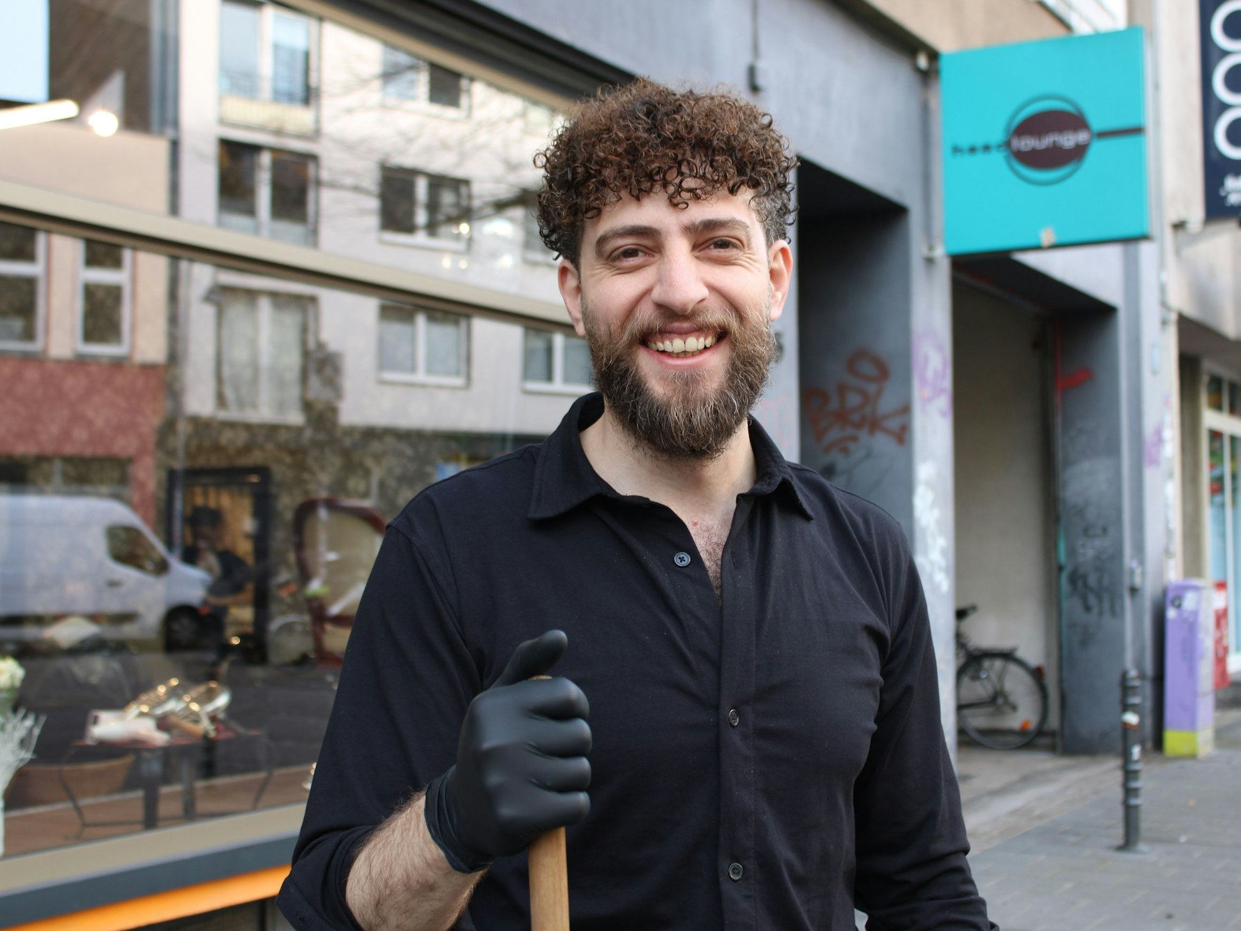 Ein Mann steht mit einem Besen und mit Gummihandschuhe vor einem Frisörsalon.