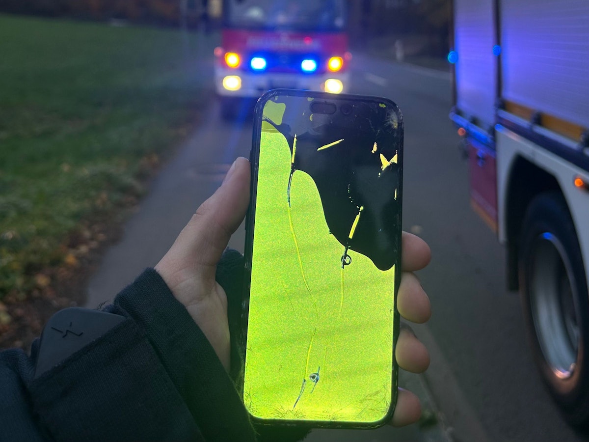 Ein Feuerwehrmann hält ein ramponiertes Handy in der Hand. Im Hintergrund sin Einsatzfahrzeuge mit Blaulicht zu sehen.