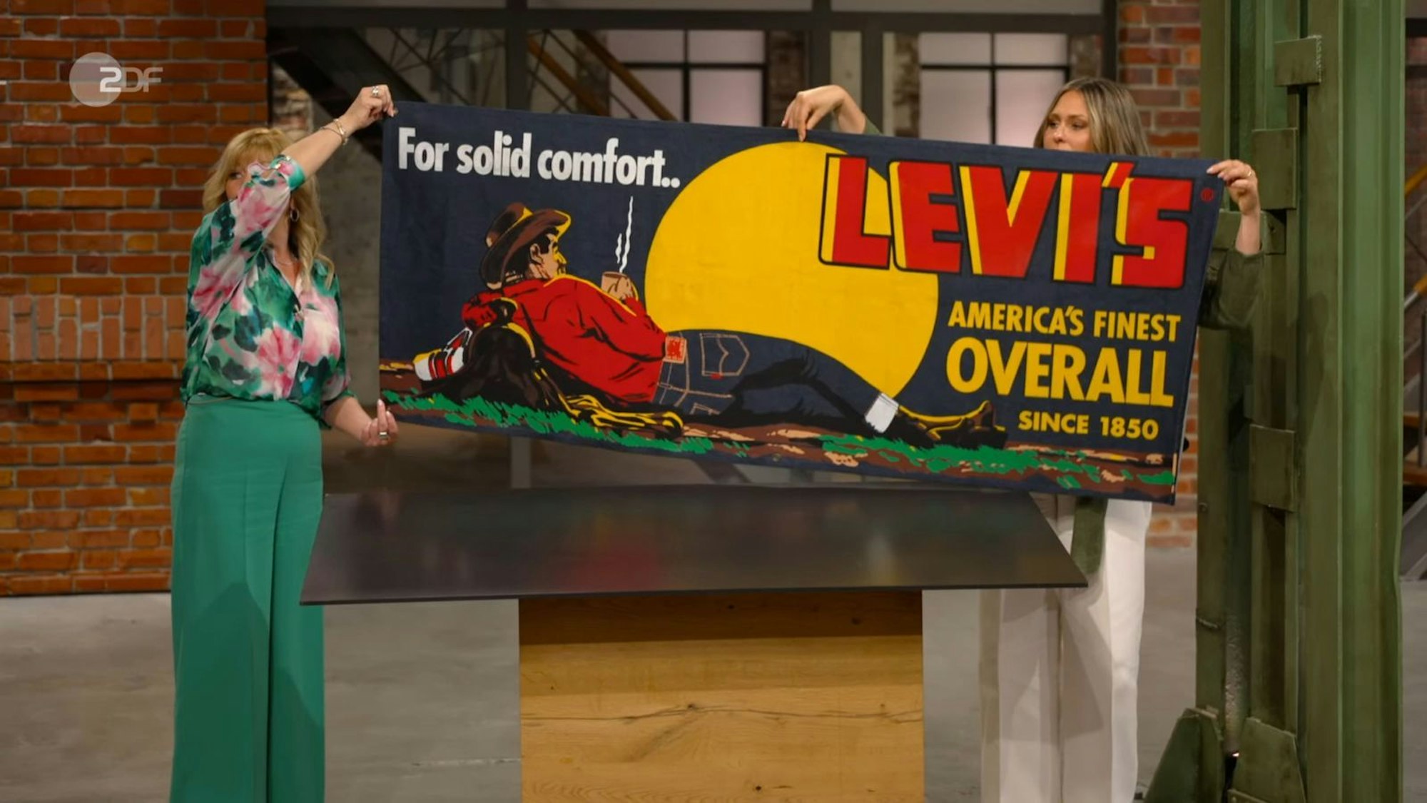 „Das ist ja cool“, schwärmte Sarah Schreiber vom wohl „stylischsten Cowboy seit langem“. Elke Velten und sie hielten für die Kollegen das Plakat hoch. „Overall?“, las Benjamin Leo Leo vor und fragte sich: „Was heißt das?“ Eine Jeans ist doch kein Overall ... (Bild: ZDF)