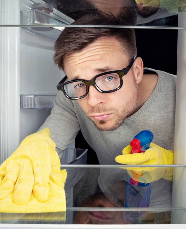 Ein Mann mit schwarzer Hornbrille und Haushaltshandschuhe guckt verdrießlich, während er einen Kühlschrank reinigt.
