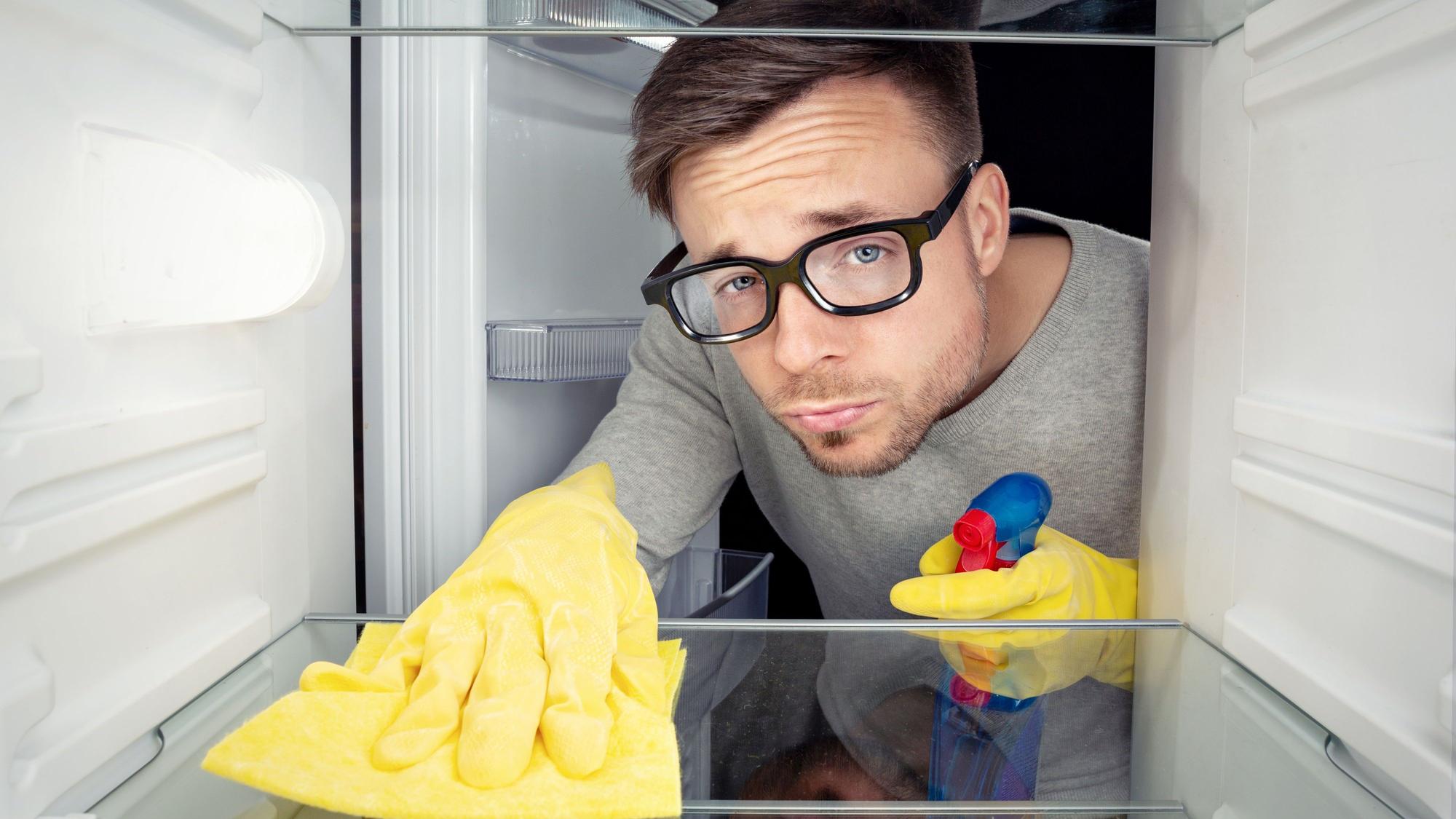 Ein Mann mit schwarzer Hornbrille und Haushaltshandschuhe guckt verdrießlich, während er einen Kühlschrank reinigt.