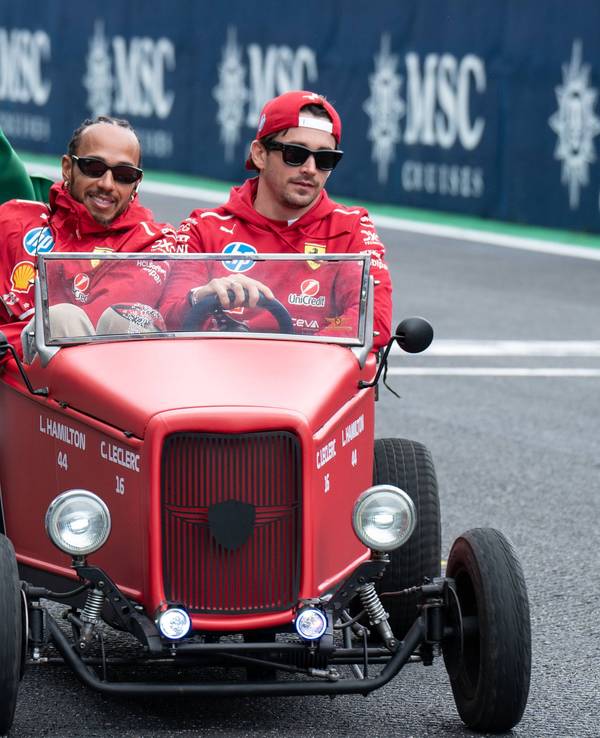 Lewis Hamilton und Charles Leclerc sitzen gemeinsam in einem kleinen alten roten Auto und fahren über die Rennstrecke in Brasilien. Hamilton hält dabei eine Brasilien-Flagge an der Seite des Autos raus, Leclerc lenkt den Wagen.
