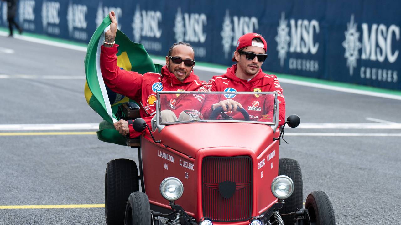 Lewis Hamilton und Charles Leclerc sitzen gemeinsam in einem kleinen alten roten Auto und fahren über die Rennstrecke in Brasilien. Hamilton hält dabei eine Brasilien-Flagge an der Seite des Autos raus, Leclerc lenkt den Wagen.