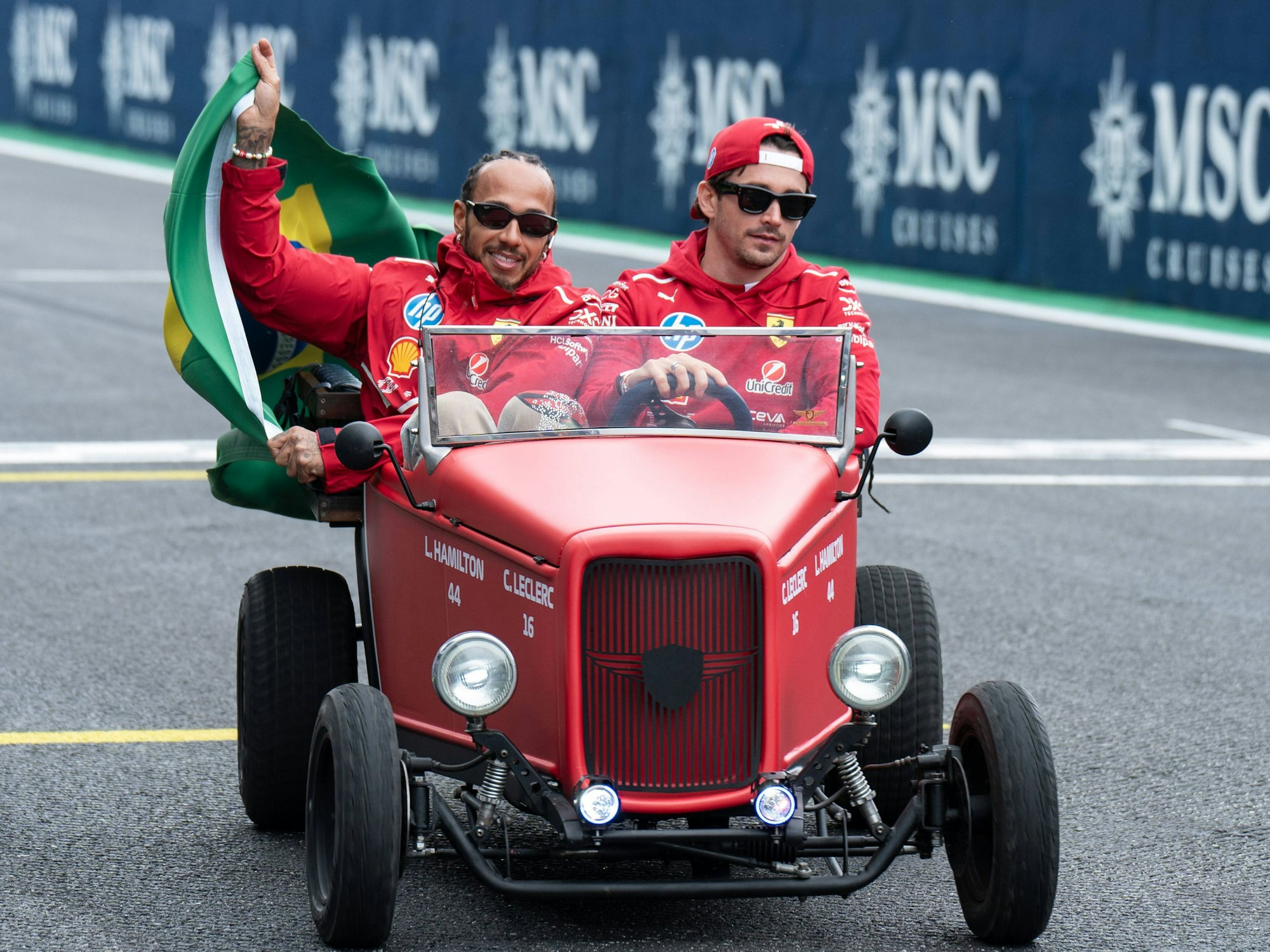 Lewis Hamilton und Charles Leclerc sitzen gemeinsam in einem kleinen alten roten Auto und fahren über die Rennstrecke in Brasilien. Hamilton hält dabei eine Brasilien-Flagge an der Seite des Autos raus, Leclerc lenkt den Wagen.
