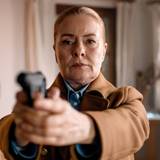 Eine Frau mittleren Alters mit streng zurückgekämmten Haaren richtet eine Pistole in Richtung Kamera.